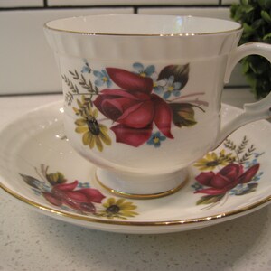 Può includere: Una tazza da tè e un piattino bianchi con un bordo dorato e un motivo floreale. Il motivo presenta rose rosse, fiori gialli e nontiscordardimé.