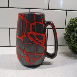 K&ouml;nnte beinhalten: Eine schwarze Keramiktasse mit einem roten, netzartigen Design. Die Tasse hat einen abgerundeten K&ouml;rper und einen Henkel.