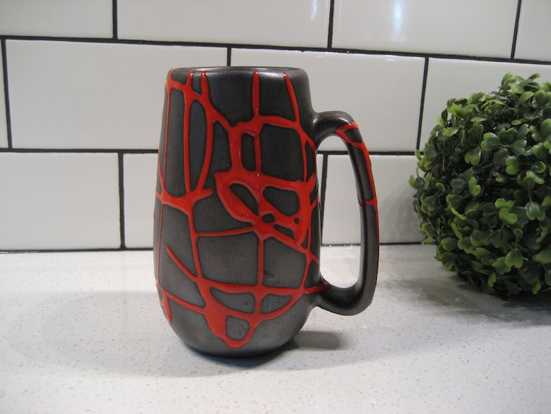 K&ouml;nnte beinhalten: Eine schwarze Keramiktasse mit einem roten, netzartigen Design. Die Tasse hat einen abgerundeten K&ouml;rper und einen Henkel.