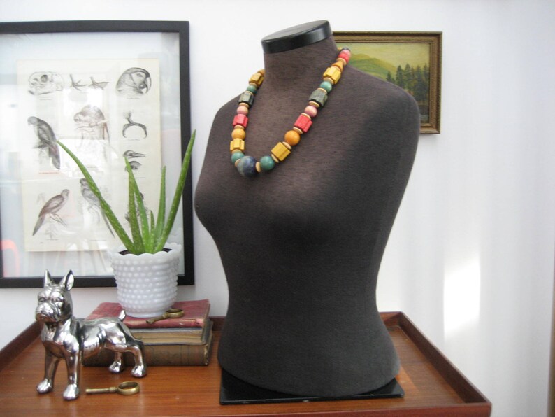May include: A black fabric mannequin with a colorful beaded necklace. The necklace has yellow, orange, red, green, and blue beads. The mannequin is on a wooden table.