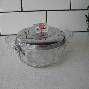 May include: A clear glass sugar bowl with a silver lid and a colorful plastic knob. The bowl has a textured design and is sitting on a white countertop.