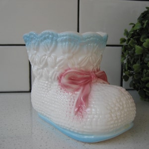 Könnte beinhalten: Ein weißer Keramik-Babyschuh-Pflanztopf mit blau-weißem Spitzenbesatz und einer rosa Schleife. Der Topf steht auf einer weißen gefliesten Oberfläche.