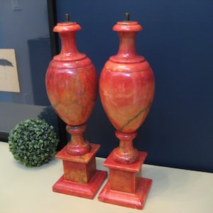 May include: Two red marble urns with a swirl pattern on a white surface. The urns are tall and slender with a round base and a flared top.
