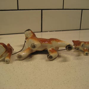 May include: Three ceramic figurines of dogs connected by a chain. The dogs are brown and white with black eyes and noses. The largest dog is in the middle and the two smaller dogs are on either side.