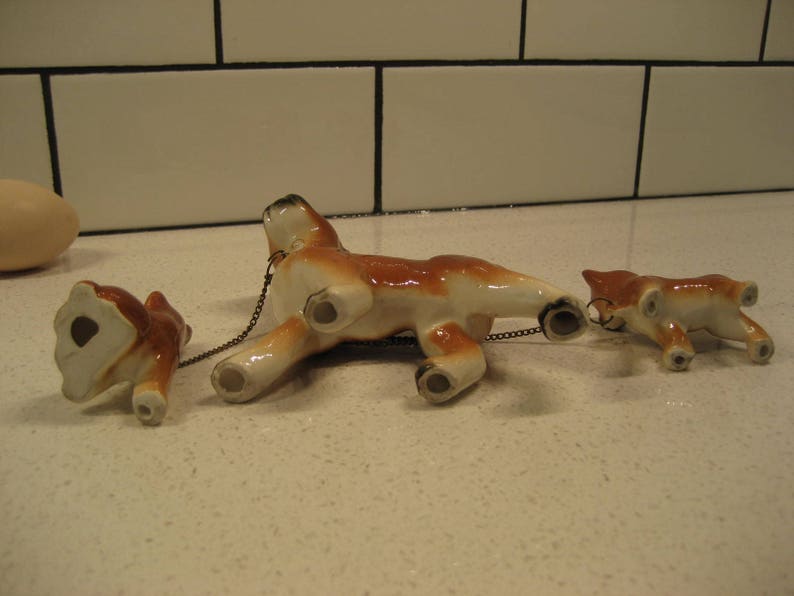May include: Three ceramic figurines of dogs connected by a chain. The dogs are brown and white with black eyes and noses. The largest dog is in the middle and the two smaller dogs are on either side.