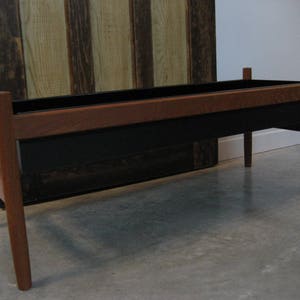 Könnte beinhalten: Ein Holz-Pflanzgefäß mit einer schwarzen Metallauskleidung. Das Pflanzgefäß ist aus dunklem Holz und hat eine rechteckige Form. Das Pflanzgefäß steht auf einem Betonboden.