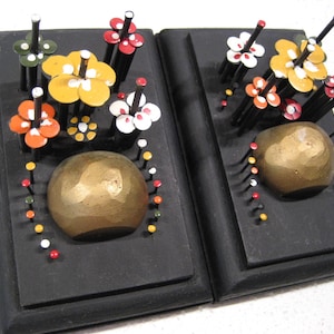 May include: Two black wooden boxes with gold round objects in the center. Each box is decorated with colorful flowers made of small nails with painted heads. The flowers are red, white, green, yellow, and orange.