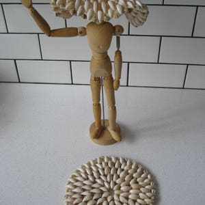 May include: A wooden artist's mannequin wearing a hat made of white seashells. The mannequin is standing on a white surface with a round, white seashell mat in front of it.