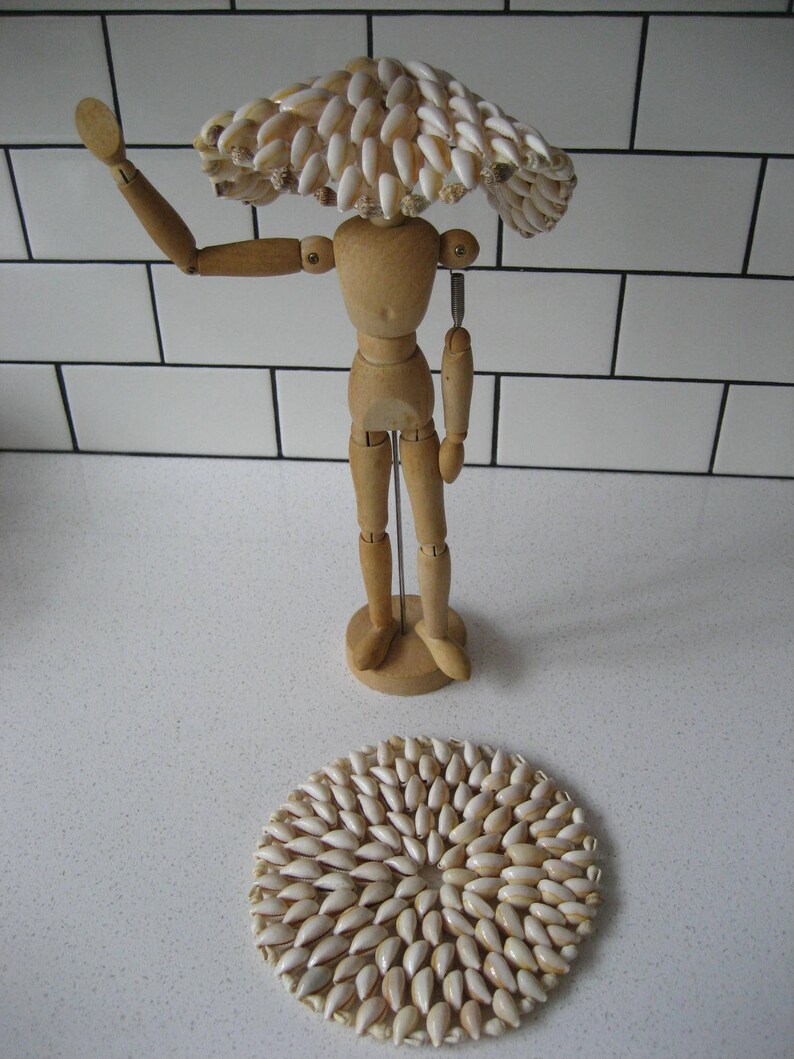 May include: A wooden artist's mannequin wearing a hat made of white seashells. The mannequin is standing on a white surface with a round, white seashell mat in front of it.