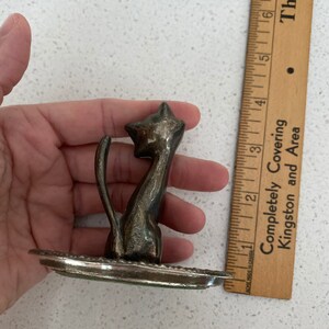 K&ouml;nnte beinhalten: Eine silberfarbene Katzenfigur aus Metall mit einem hochgestellten Schwanz und einem dekorativen Sockel. Die Katze sitzt aufrecht mit dem Kopf zur Seite geneigt.