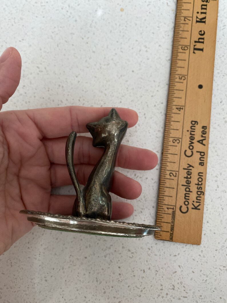 K&ouml;nnte beinhalten: Eine silberfarbene Katzenfigur aus Metall mit einem hochgestellten Schwanz und einem dekorativen Sockel. Die Katze sitzt aufrecht mit dem Kopf zur Seite geneigt.