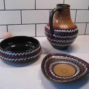 以下が含まれることがあります： 黒と白の幾何学模様と赤いアクセントが施された、セラミック製の食器3点セット。食器は、ボウル、プレート、ピッチャーの形をしています。