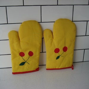 Puede incluir: Un par de guantes de horno amarillos con cerezas rojas y hojas verdes bordadas. Los guantes tienen un borde rojo.