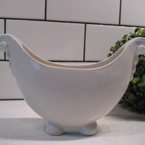 May include: A white ceramic planter shaped like a boat with a curved handle on each end. The planter has a glossy finish and sits on four small feet.