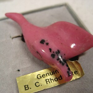 May include: A pink bird carving made of rhodonite with black markings. The carving is displayed on a white background with a gold label that reads "Genuine B.C. Rhodonite".