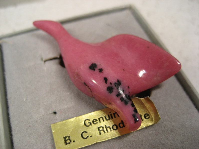 May include: A pink bird carving made of rhodonite with black markings. The carving is displayed on a white background with a gold label that reads "Genuine B.C. Rhodonite".