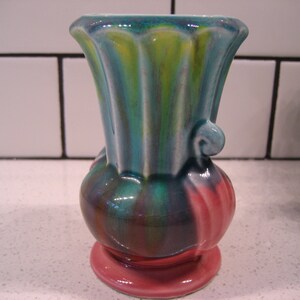 May include: A small, ceramic vase with a green, blue and pink swirl pattern. The vase has a flared top and a rounded base.