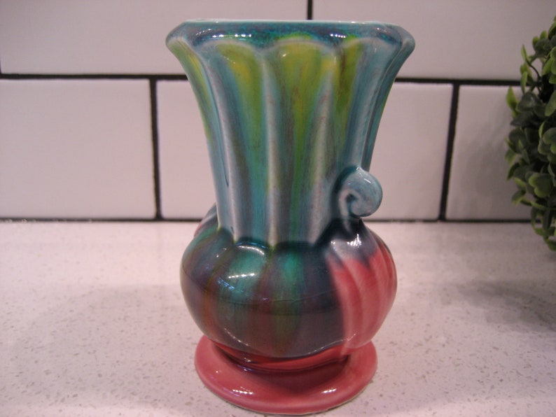 May include: A small, ceramic vase with a green, blue and pink swirl pattern. The vase has a flared top and a rounded base.