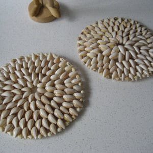 May include: Two round coasters made of white seashells arranged in a circular pattern. The coasters are on a white surface.