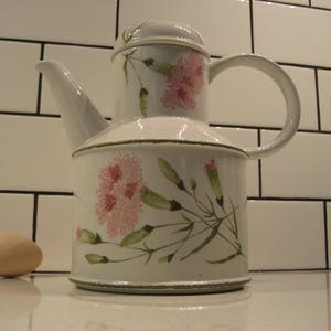 May include: A white ceramic teapot with a lid and a curved handle. The teapot is decorated with pink floral designs and green leaves. A brown egg sits to the left of the teapot. The teapot is approximately 8 inches tall.