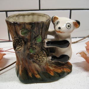 May include: A ceramic planter shaped like a tree trunk with a panda bear climbing up the side. The planter is brown, green, and white with black accents. The panda bear is white with black eyes, nose, and paws.
