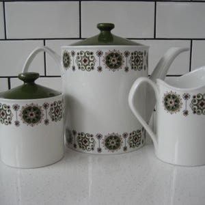 Puede incluir: Un juego de tres teteras de cerámica blanca con diseños florales verdes y marrones. La tetera tiene una tapa y un pico. La jarra de crema tiene un asa. El azucarero tiene una tapa.