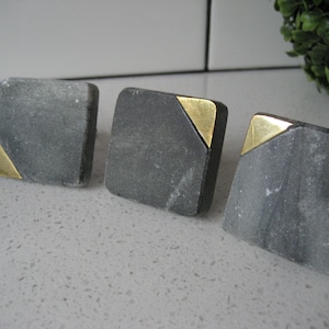 May include: Four square black marble cabinet knobs with gold accents. The knobs have a geometric design with a gold triangle on each corner.
