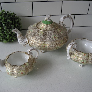 Può includere: Un set da tè vintage con teiera, lattiera e zuccheriera. Il set è decorato con un motivo floreale in tonalità di verde, oro e bianco.