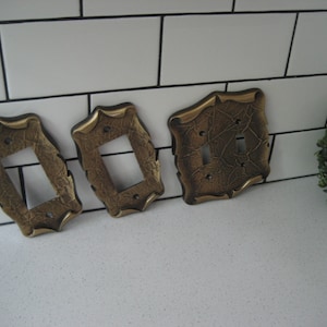 May include: Three decorative light switch covers in a gold finish with an ornate design. The covers are on a white tiled surface.