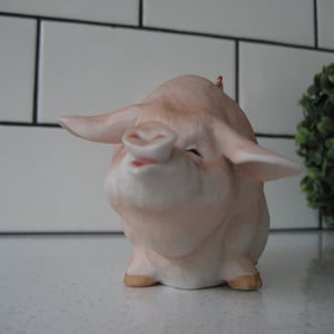 May include: A ceramic pig figurine with a smiling expression. The pig is primarily light pink with darker pink shading and tan hooves. The ears are large and spread out. The figurine is on a white surface.