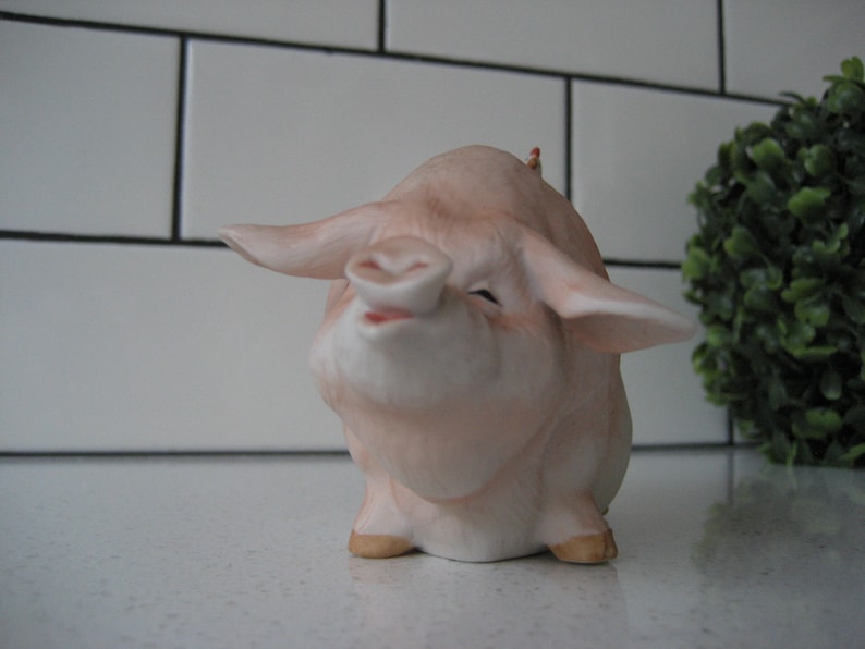 May include: A ceramic pig figurine with a smiling expression. The pig is primarily light pink with darker pink shading and tan hooves. The ears are large and spread out. The figurine is on a white surface.