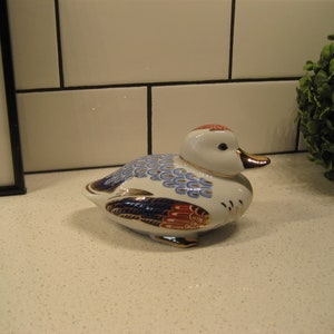 Op de afbeelding: Een wit porseleinen eendenbeeldje met blauwe, rode en gouden beschilderde details. De eend heeft een rond lichaam en een lange, dunne snavel.