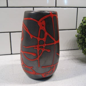 K&ouml;nnte beinhalten: Eine schwarze Keramikvase mit einem roten abstrakten Design. Die Vase hat eine abgerundete Form und eine schmale &Ouml;ffnung.