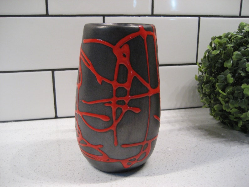 K&ouml;nnte beinhalten: Eine schwarze Keramikvase mit einem roten abstrakten Design. Die Vase hat eine abgerundete Form und eine schmale &Ouml;ffnung.