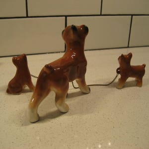 May include: Three ceramic figurines of brown and white boxer dogs. The largest dog is standing and the two smaller dogs are sitting. The dogs are connected by a chain.