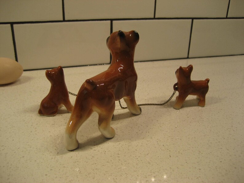 May include: Three ceramic figurines of brown and white boxer dogs. The largest dog is standing and the two smaller dogs are sitting. The dogs are connected by a chain.