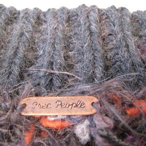May include: A close-up of a gray and brown knitted scarf with a copper tag that reads "Free People".