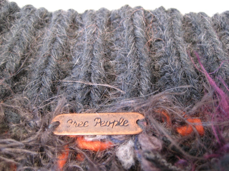 May include: A close-up of a gray and brown knitted scarf with a copper tag that reads "Free People".