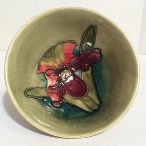 Puede incluir: Un cuenco de cerámica verde con un diseño floral en relieve. La flor es una orquídea roja, rosa y amarilla con hojas verdes.