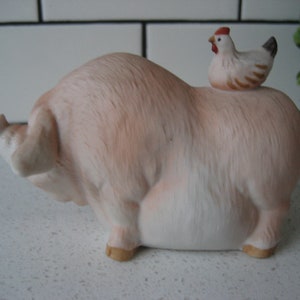 May include: A ceramic figurine of a light pink pig with a small white and brown chicken perched on its back. The pig has a detailed, textured surface and is in a resting position. The chicken has a red comb and a black tail.