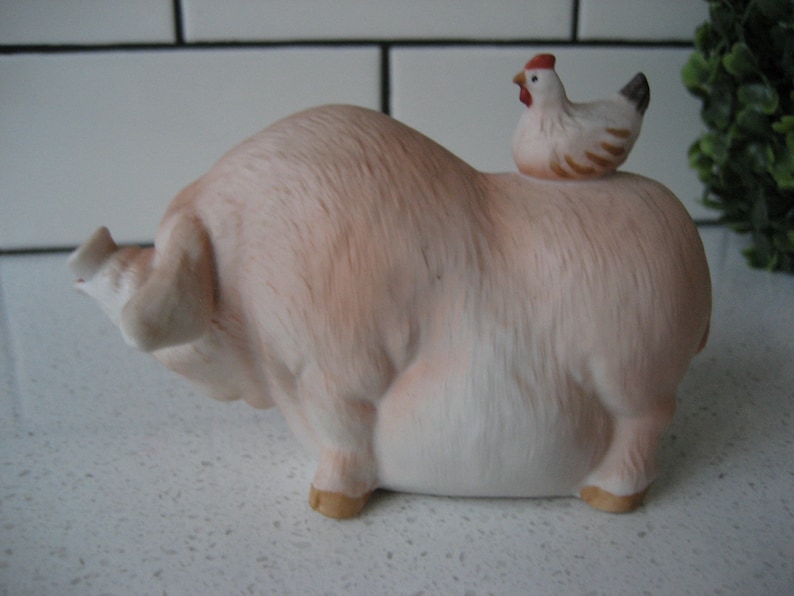 May include: A ceramic figurine of a light pink pig with a small white and brown chicken perched on its back. The pig has a detailed, textured surface and is in a resting position. The chicken has a red comb and a black tail.