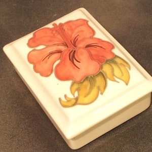 Puede incluir: Una caja de cerámica blanca con un diseño en relieve de una flor de hibisco rosa con hojas amarillas.