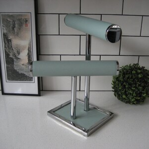 May include: A chrome jewelry stand with two light blue padded arms for displaying bracelets and necklaces. The stand has a square base with a light blue padded surface.