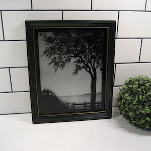 Może przedstawiać: Czarno-biała sylwetka przedstawiająca dom, drzewo i płot nad jeziorem. Dzieło jest oprawione w czarną ramę ze złotą ozdobą.