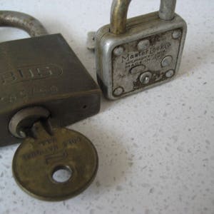 May include: Two vintage padlocks, one rectangular and one square, sit side-by-side. The rectangular lock is dark brown with the word "ABUS" and "N 85/50" printed on it. A brass key is in the keyhole. The square lock is silver with the words "Master Lock" printed on it.
