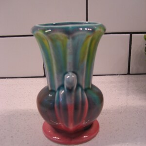 May include: A small, pink and blue ceramic vase with a unique, abstract design. The vase has a wide, round base and a narrow, cylindrical neck.