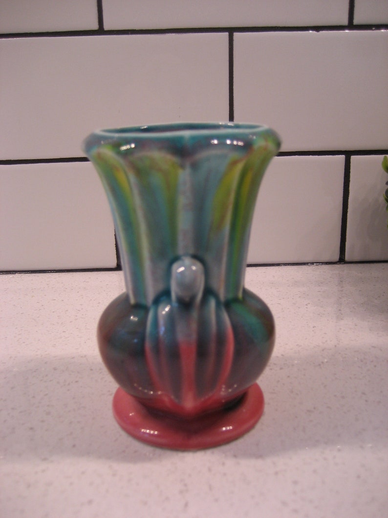 May include: A small, pink and blue ceramic vase with a unique, abstract design. The vase has a wide, round base and a narrow, cylindrical neck.