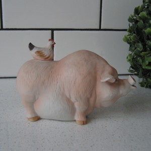 May include: A ceramic figurine of a light pink pig with a small chicken perched on its back. The pig has a detailed snout and small hooves. The chicken is white, brown, and black with a red comb.