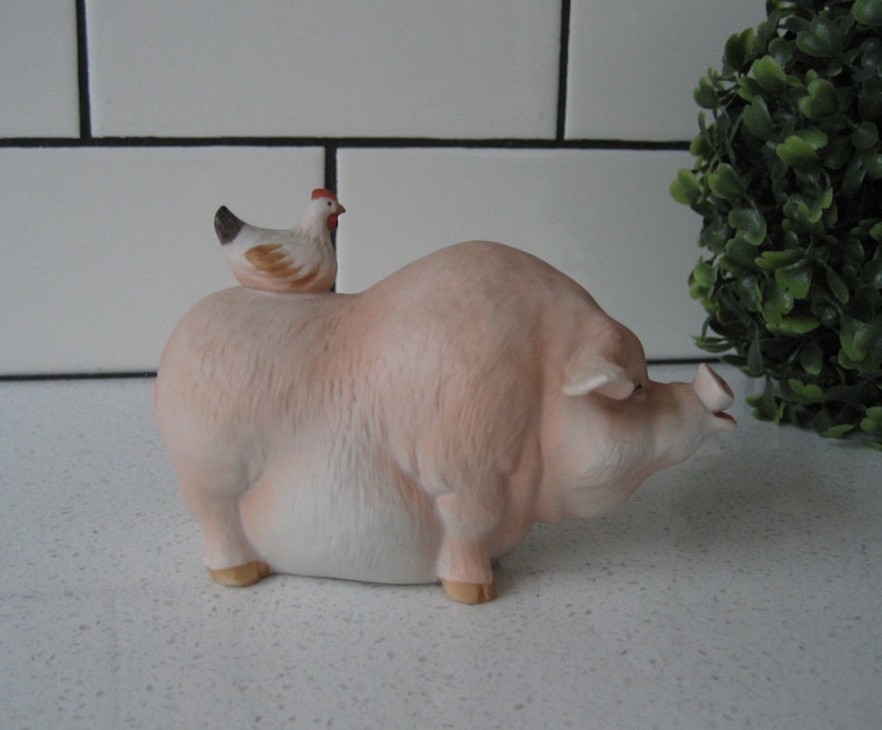 May include: A ceramic figurine of a light pink pig with a small chicken perched on its back. The pig has a detailed snout and small hooves. The chicken is white, brown, and black with a red comb.
