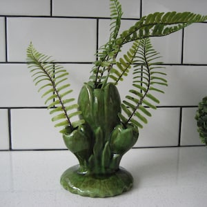 Op de afbeelding: Een groene keramische vaas met drie openingen voor bloemen. De vaas is gevormd als een gestileerde bloem met een brede basis en een smalle hals. De vaas is versierd met een groene glazuur en heeft een getextureerd oppervlak. De vaas is gevuld met groene varens.
